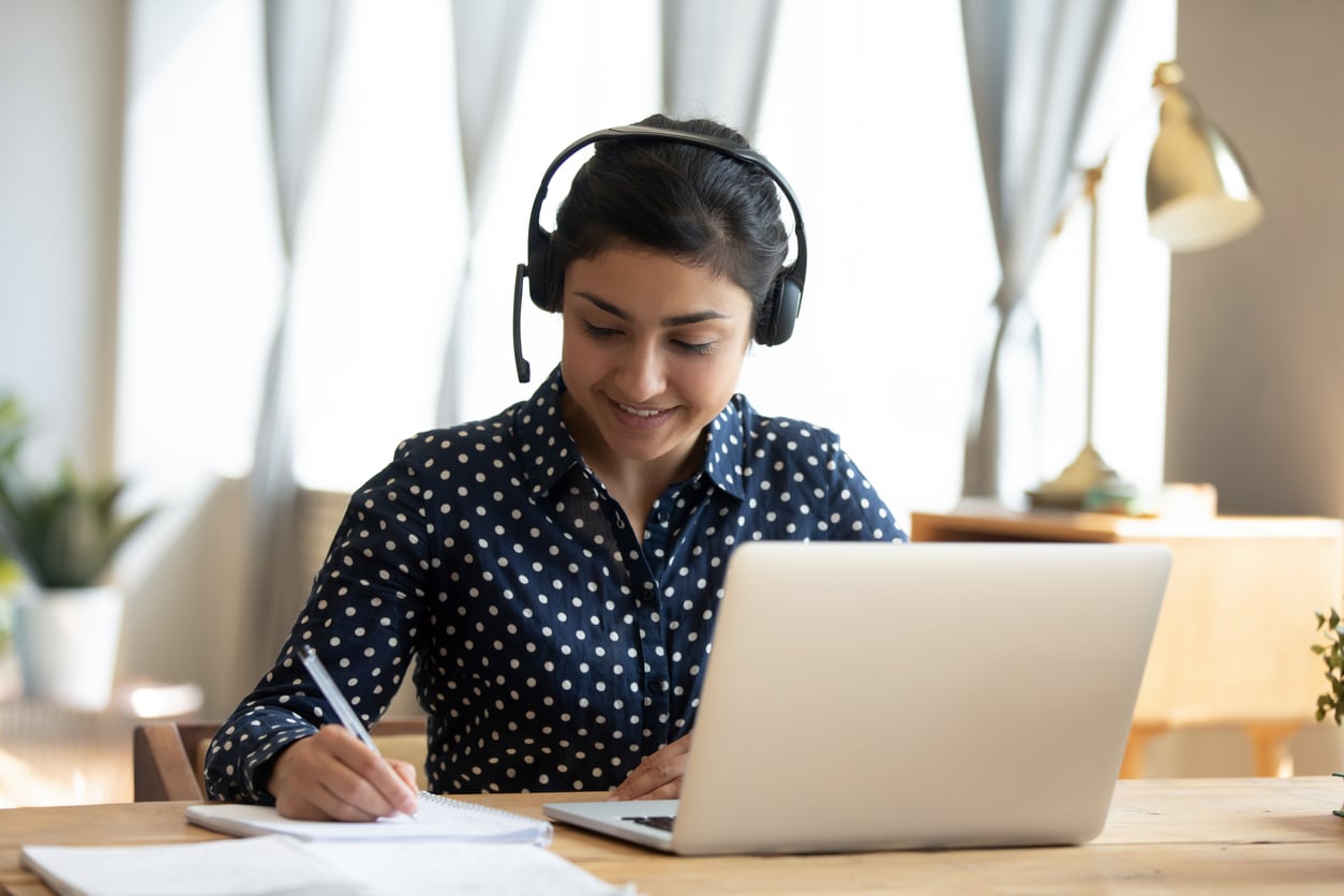 A young woman wearing headphones, writing notes while working on a laptop at a bright desk.
